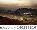 Sunlight streams through dark clouds, illuminating parts of a valley and casting shadows on distant mountain ranges. 140233816