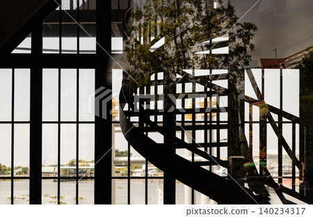 An industrial black spiral staircase serves as a sweeping focal point, winding upward past towering gridded windows. Through the glass, a serene waterfront stretches into the distance beneath a bright 140234317