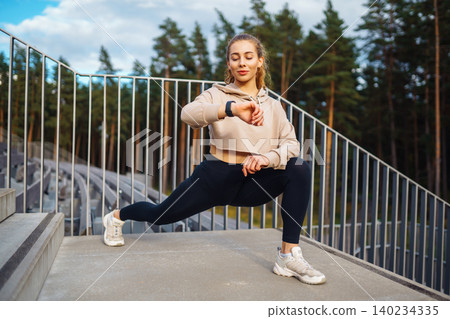 Active woman in sportswear does stretching exercise, looks at fitness application through Smartwatch Active woman in sportswear does stretching exercise, looks at fitness application through Smartwatch 140234335