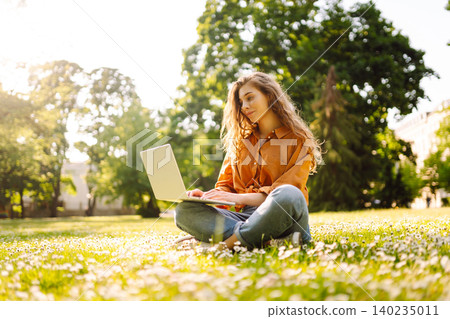 Woman in casual clothes with a laptop sits on lawn on sunny day. Happy freelancer working outdoors. Woman in casual clothes with a laptop sits on lawn on sunny day. Happy freelancer working outdoors. 140235011