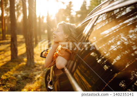 A young woman feels freedom leaning out of a car window in a sunny forest. Travel concept. A young woman feels freedom leaning out of a car window in a sunny forest. Travel concept. 140235141