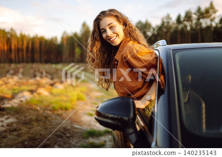A young woman feels freedom leaning out of a car window in a sunny forest. Travel concept. A young woman feels freedom leaning out of a car window in a sunny forest. Travel concept. 140235143
