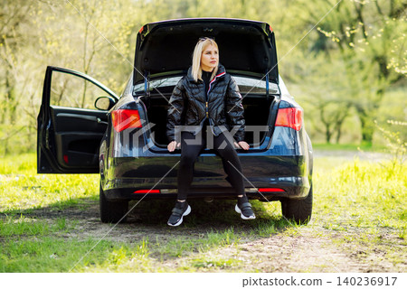 fair haired woman in a black jacket sits in the open trunk of a blue automobile in a clearing 140236917