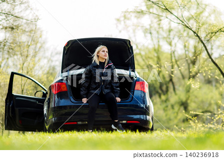 woman in dark clothing sits in the open trunk of a blue car against the background of trees from below 140236918