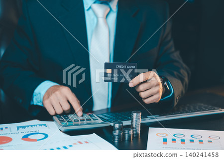 Close up of businessman hand holding a black credit card and using calculator with coins stack on table. Financial planning concept. Close up of businessman hand holding a black credit card and using calculator with coins stack on table. Financial planning concept. 140241458