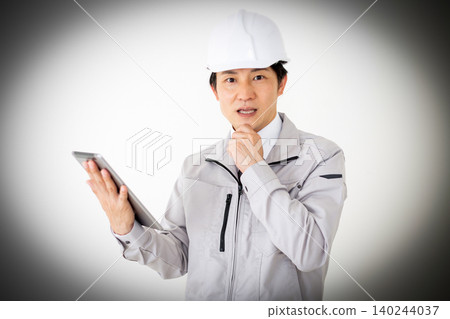 一位身穿灰色工作服、頭戴安全帽的中年商務人士正看著平板電腦,臉上露出驚訝的表情。 (白色背景) 一位身穿灰色工作服、頭戴安全帽的中年商務人士正看著平板電腦,臉上露出驚訝的表情。 (白色背景) 140244037