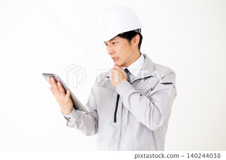 一位身穿灰色工作服、頭戴安全帽的中年商人,在白色背景前看著平板電腦,若有所思。 一位身穿灰色工作服、頭戴安全帽的中年商人,在白色背景前看著平板電腦,若有所思。 140244038