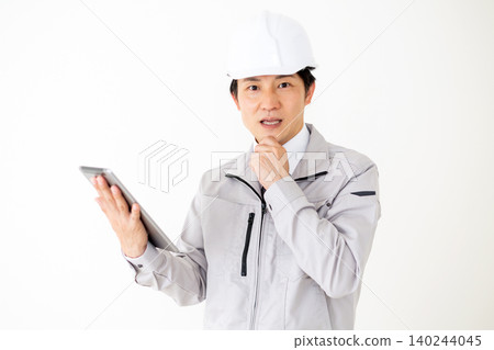 A middle-aged businessman in a gray work uniform and helmet looks surprised while looking at a tablet. (White background) 140244045