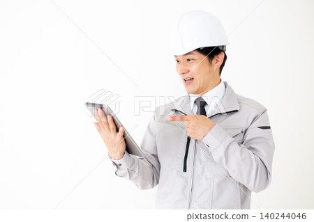 一位身穿灰色工作服、頭戴安全帽的中年商務人士正看著平板電腦,臉上露出驚訝的表情。 (白色背景) 一位身穿灰色工作服、頭戴安全帽的中年商務人士正看著平板電腦,臉上露出驚訝的表情。 (白色背景) 140244046