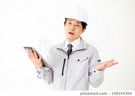 A middle-aged businessman in a gray work uniform and helmet, looking at a tablet and throwing his hands up in frustration, against a white background. A middle-aged businessman in a gray work uniform and helmet, looking at a tablet and throwing his hands up in frustration, against a white background. 140244364