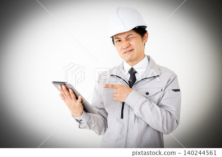 A middle-aged businessman in gray work clothes and a helmet, looking thoughtfully at a tablet, against a white background. A middle-aged businessman in gray work clothes and a helmet, looking thoughtfully at a tablet, against a white background. 140244561