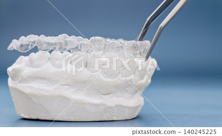 Clear invisible aligners held by tweezers over a plaster teeth model on a blue background. Orthodontic concept. Clear invisible aligners held by tweezers over a plaster teeth model on a blue background. Orthodontic concept. 140245225