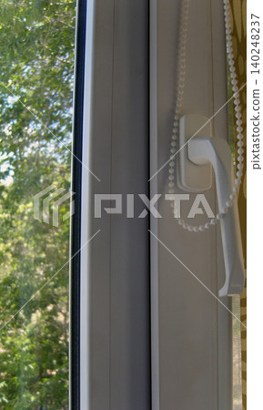 Modern white uPVC window. The focus is on the frame, the ergonomic handle, and the beaded pull chain for a pull-down blind. The handle is positioned for right-hand use Modern white uPVC window. The focus is on the frame, the ergonomic handle, and the beaded pull chain for a pull-down blind. The handle is positioned for right-hand use 140248237