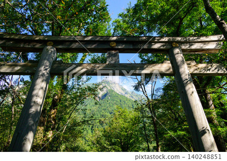 A wooden torii gate rising above, and the beautiful summer mountain scenery seen through the lush green forest. A wooden torii gate rising above, and the beautiful summer mountain scenery seen through the lush green forest. 140248851