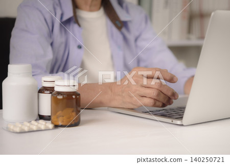 Close up senior hand using laptop with medicine bottles. Elderly patient ordering prescription drug from online pharmacy, researching vitamin supplement or consulting doctor via telemedicine at home. Close up senior hand using laptop with medicine bottles. Elderly patient ordering prescription drug from online pharmacy, researching vitamin supplement or consulting doctor via telemedicine at home. 140250721