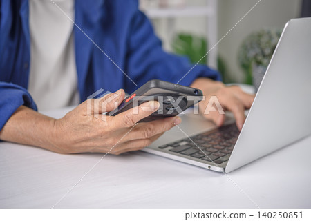 Close up of senior woman hands holding smartphone and typing on laptop keyboard. Elderly businesswoman multitasking with technology, data entry, and digital connection concept. Close up of senior woman hands holding smartphone and typing on laptop keyboard. Elderly businesswoman multitasking with technology, data entry, and digital connection concept. 140250851