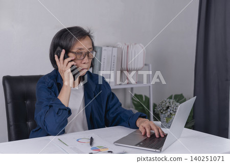 Senior Asian business woman talking on smartphone and working on laptop with financial charts at home office. Elderly entrepreneur multitasking, remote work, and business consulting concept. 140251071