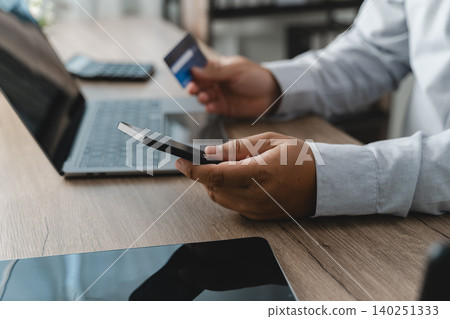 Person holding a smartphone and credit card while using a laptop for mobile banking, online payment authorization and digital financial transaction management in a cashless environment. Person holding a smartphone and credit card while using a laptop for mobile banking, online payment authorization and digital financial transaction management in a cashless environment. 140251333