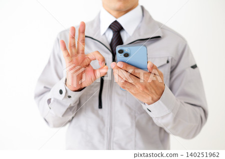 A middle-aged businessman in gray work clothes makes a circular sign with his smartphone against a white background. A middle-aged businessman in gray work clothes makes a circular sign with his smartphone against a white background. 140251962