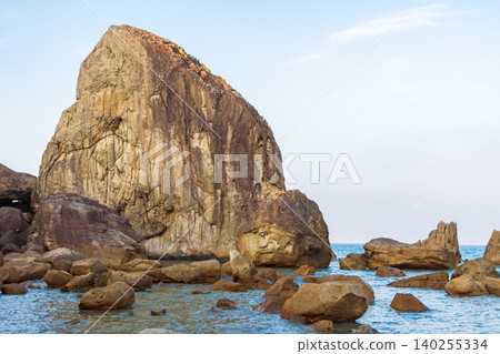 A giant, strangely shaped rock jutting out of the sea against a backdrop of blue sky and the rocky shoreline. A giant, strangely shaped rock jutting out of the sea against a backdrop of blue sky and the rocky shoreline. 140255334