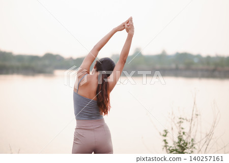 Rear view of young woman doing outdoor yoga back stretch with raised arms by a calm lakeside at sunrise, expressing wellness, freedom, mindfulness, relaxation, nature fitness, and peace. Rear view of young woman doing outdoor yoga back stretch with raised arms by a calm lakeside at sunrise, expressing wellness, freedom, mindfulness, relaxation, nature fitness, and peace. 140257161