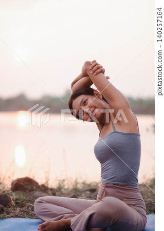 Young woman doing seated yoga stretch on a mat beside a calm lakeside at sunrise, showing flexibility, wellness, mindfulness, mobility, relaxation, healthy lifestyle, and nature fitness. Young woman doing seated yoga stretch on a mat beside a calm lakeside at sunrise, showing flexibility, wellness, mindfulness, mobility, relaxation, healthy lifestyle, and nature fitness. 140257164