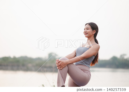 Asian woman holding standing knee stretch beside a calm lake, showing balance, flexibility, mobility, outdoor wellness, mindful breathing, recovery, posture, fitness routine, and relaxation. Asian woman holding standing knee stretch beside a calm lake, showing balance, flexibility, mobility, outdoor wellness, mindful breathing, recovery, posture, fitness routine, and relaxation. 140257339