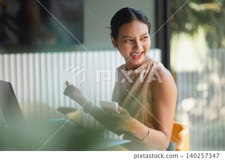 Cheerful Asian businesswoman holding smartphone beside laptop in modern cafe office, smiling during client communication, financial planning, online work, accounting, productivity, and success. Cheerful Asian businesswoman holding smartphone beside laptop in modern cafe office, smiling during client communication, financial planning, online work, accounting, productivity, and success. 140257347