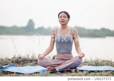 Asian woman meditating in lotus pose on yoga mat beside a calm lake, showing mindfulness, breathing, peaceful nature, wellness lifestyle, relaxation, balance, mental focus, and serenity. 140257373