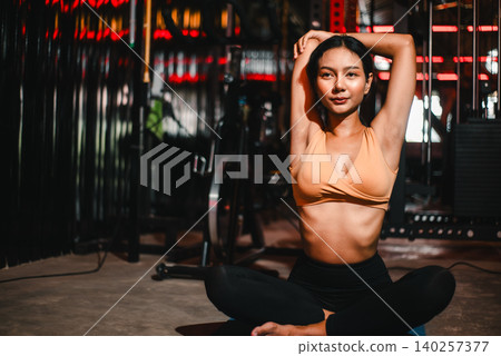 Asian woman doing seated overhead arm stretch on yoga mat in a dark gym, showing flexibility, mobility, wellness, recovery, mindfulness, fitness routine, posture, strength, and relaxed focus. 140257377