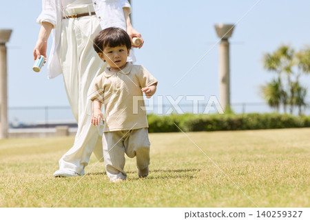 一個蹣跚學步的孩子在草地公園裡走來走去,他的母親在一旁照顧。 一個蹣跚學步的孩子在草地公園裡走來走去,他的母親在一旁照顧。 140259327