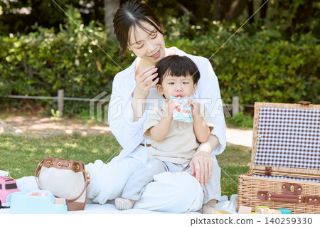 A mother enjoying a picnic in the park with her toddler eating a baby food pouch. A mother enjoying a picnic in the park with her toddler eating a baby food pouch. 140259330