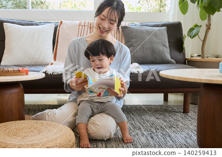一位母親在客廳和她笑容燦爛的幼兒一起讀圖畫書。 140259413