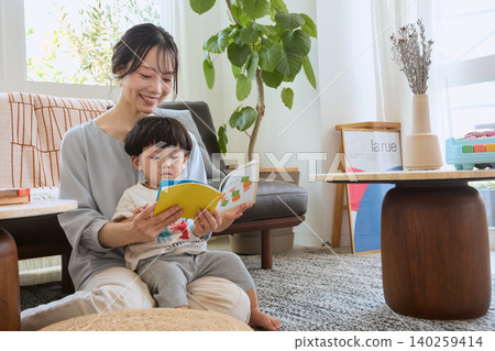 一位年輕的母親在客廳一邊為孩子讀圖畫書，一邊露出笑容。 140259414
