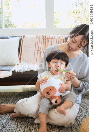 A mother watches over a toddler brushing their teeth and a baby holding a stuffed animal in the living room. 140259421