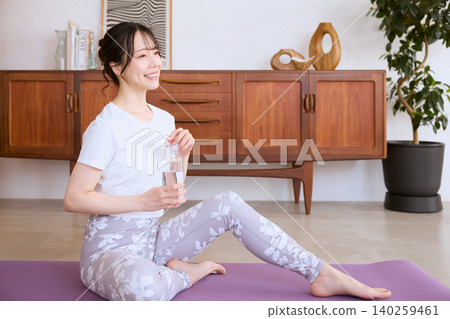 一位微笑的女子在家中做瑜珈休息時喝水。 一位微笑的女子在家中做瑜珈休息時喝水。 140259461