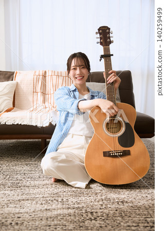 A smiling young woman playing an acoustic guitar on the living room sofa. A smiling young woman playing an acoustic guitar on the living room sofa. 140259499