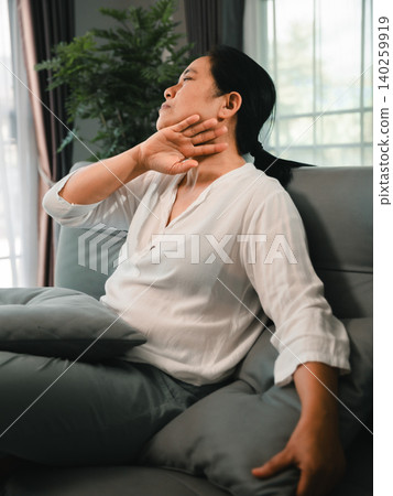 Asian woman sitting on a sofa at home with neck discomfort, stretching her shoulder and throat area, representing muscle tension, fatigue, stress relief, wellness, and body care routine. Asian woman sitting on a sofa at home with neck discomfort, stretching her shoulder and throat area, representing muscle tension, fatigue, stress relief, wellness, and body care routine. 140259919