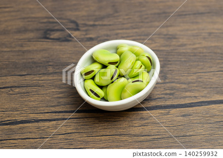 Raw broad beans in a small white bowl; wood-grain background and copy space. Raw broad beans in a small white bowl; wood-grain background and copy space. 140259932