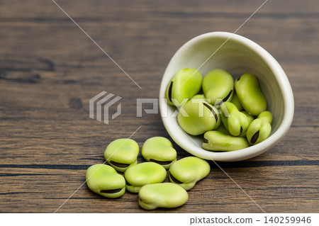 A stock photo of raw broad beans spilling from a small white plate, against a dark wood grain background. A stock photo of raw broad beans spilling from a small white plate, against a dark wood grain background. 140259946