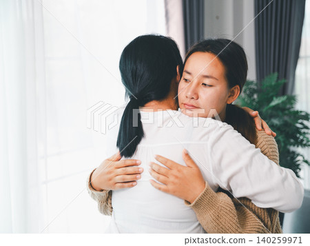Asian parent hugging a young woman at home, showing family comfort, emotional support, empathy, reconciliation, mental wellness, caregiving, trust, and a healing relationship in a bright living room. Asian parent hugging a young woman at home, showing family comfort, emotional support, empathy, reconciliation, mental wellness, caregiving, trust, and a healing relationship in a bright living room. 140259971
