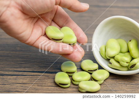 一小碟白盤子裡溢出幾顆生蠶豆,一個男人正把它們撿起來。 一小碟白盤子裡溢出幾顆生蠶豆,一個男人正把它們撿起來。 140259992