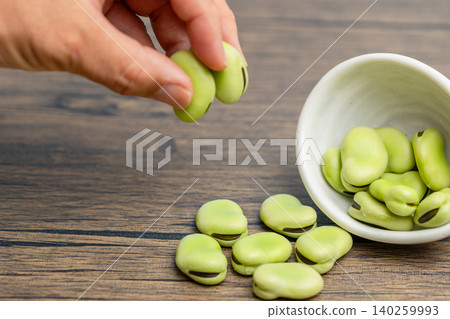 Raw broad beans spilling from a small white plate, and a man picking them up. Raw broad beans spilling from a small white plate, and a man picking them up. 140259993