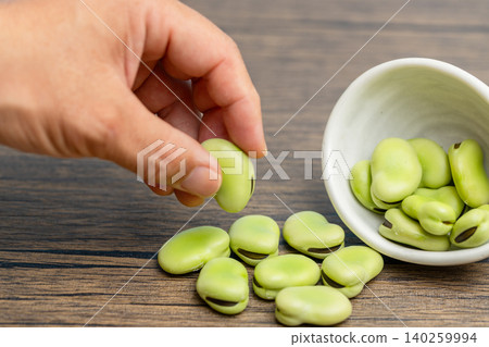 一小碟白盤子裡溢出幾顆生蠶豆,一個男人正把它們撿起來。 一小碟白盤子裡溢出幾顆生蠶豆,一個男人正把它們撿起來。 140259994