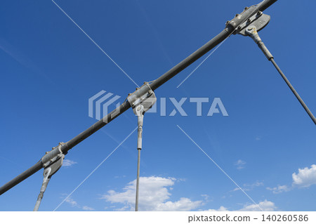 A close-up view shows metal cables and supports against a bright blue sky with a few clouds. A close-up view shows metal cables and supports against a bright blue sky with a few clouds. 140260586