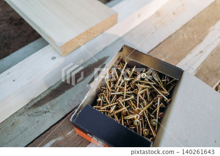 A box of construction screws rests on a larch wood board prepared for assembling a tall raised garden bed. Top view A box of construction screws rests on a larch wood board prepared for assembling a tall raised garden bed. Top view 140261643