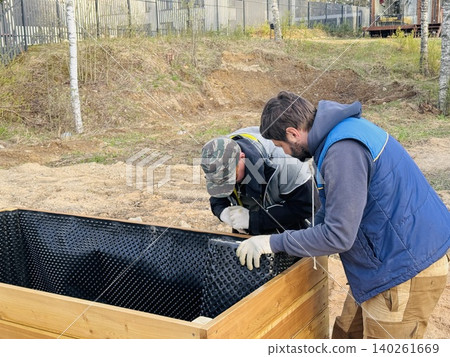 Handy men construct a tall raised garden bed from larch wood and line the inside with a protective membrane on countryside property 140261669