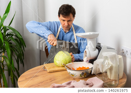 A 53-year-old man cuts cabbage with a knife while preparing to ferment cabbage and carrots. He stands in his kitchen near a vegetable grinder and a bowl of grated carrots A 53-year-old man cuts cabbage with a knife while preparing to ferment cabbage and carrots. He stands in his kitchen near a vegetable grinder and a bowl of grated carrots 140261975