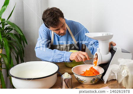 A 53-year-old man is in his kitchen preparing cabbage and carrots for fermentation. He uses a machine to shred the vegetables before placing them in a bowl A 53-year-old man is in his kitchen preparing cabbage and carrots for fermentation. He uses a machine to shred the vegetables before placing them in a bowl 140261978