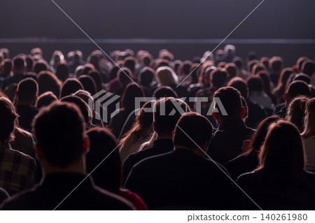 Audience watching live presentation in dark auditorium, crowd seated, silhouettes, stage lighting Audience watching live presentation in dark auditorium, crowd seated, silhouettes, stage lighting 140261980